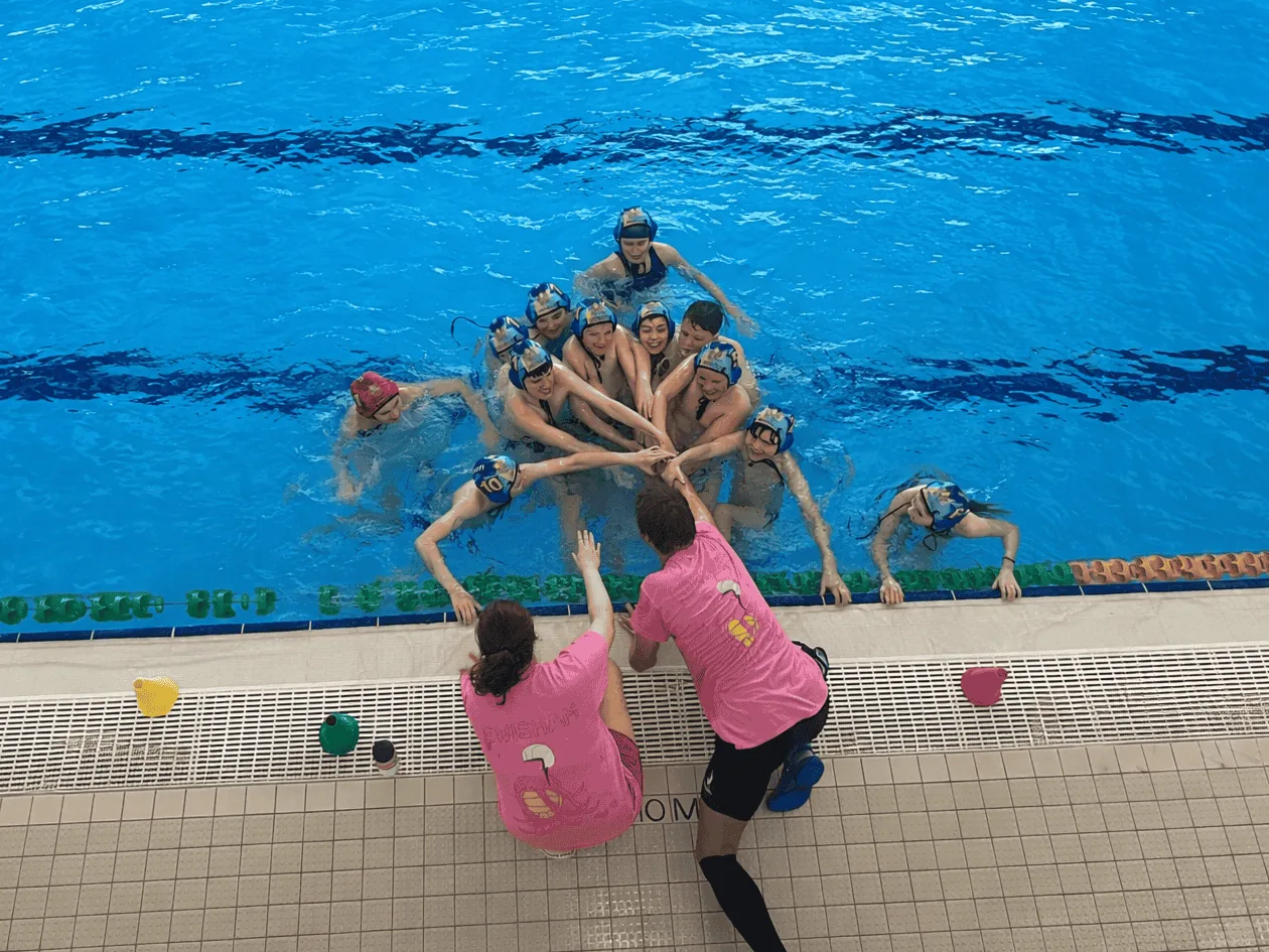 A water polo squad in a huddle at the wall, hands in the middle, two coaches crouched on the deck.